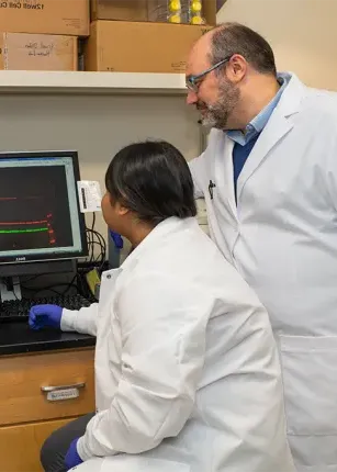 Harry Filippakis, on the left, in a while coat look at a computer screen with two graduate students in lab coats