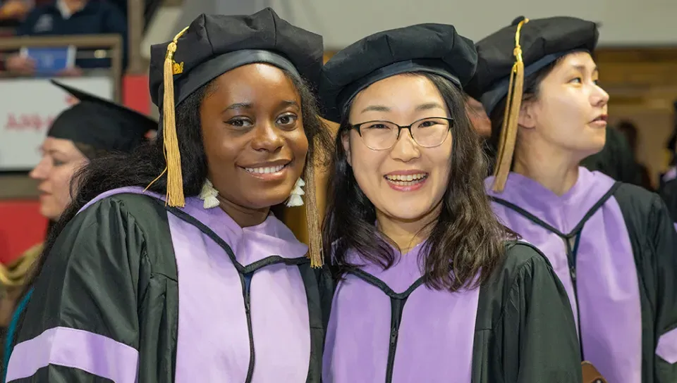 Two U N E graduate students smiling for the camera while other graduates are visible in the background