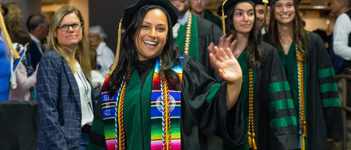 A group of U N E graduate students in black and green wave at the camera