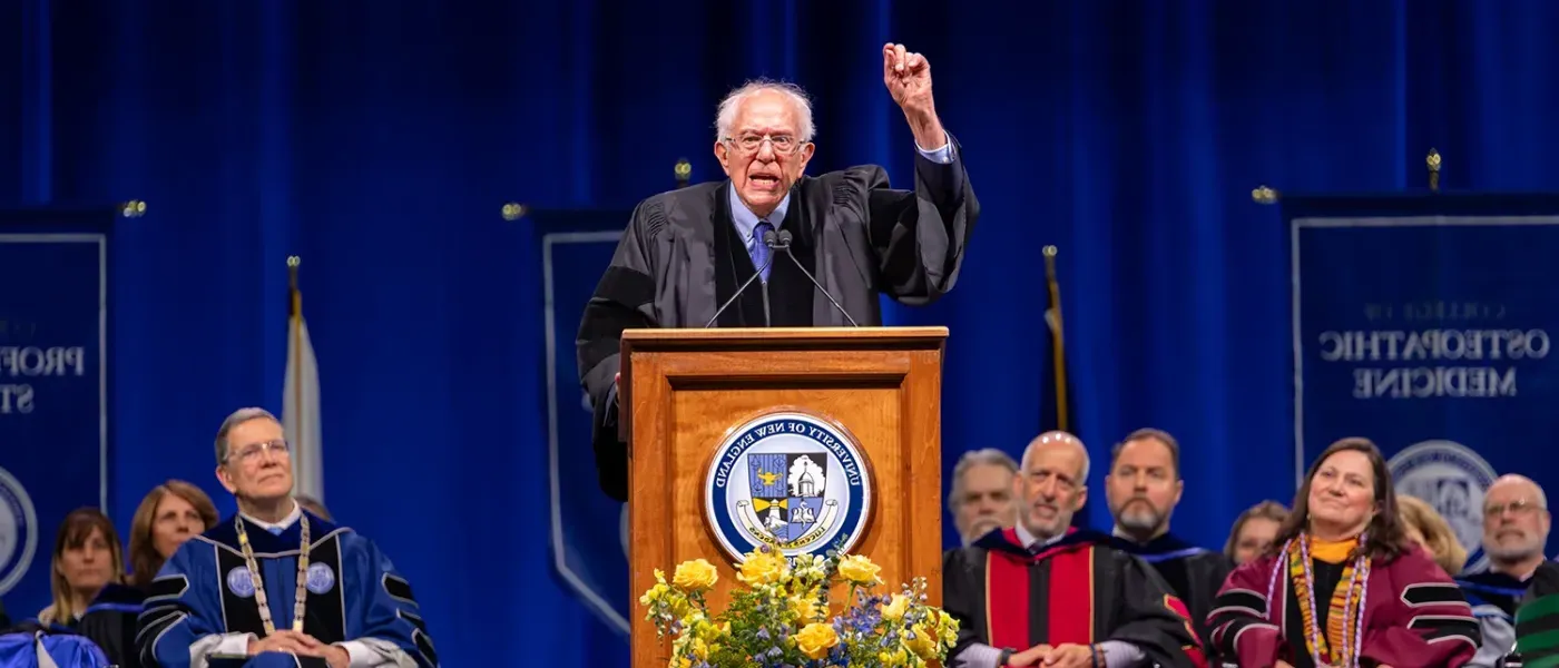 Bernie Sanders standing at the podium raises a hand gesturing to the crowd at the UNE 2024 commencement ceremony